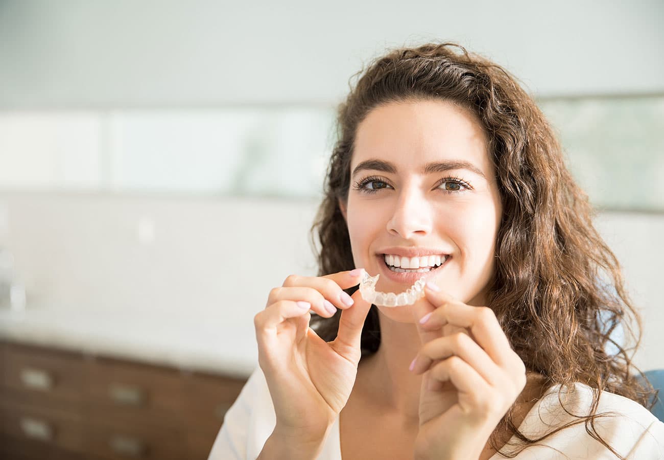 A smiling woman with wavy brown hair holds a clear aligner near her mouth, preparing to place it on her teeth in a bright, modern dental office.